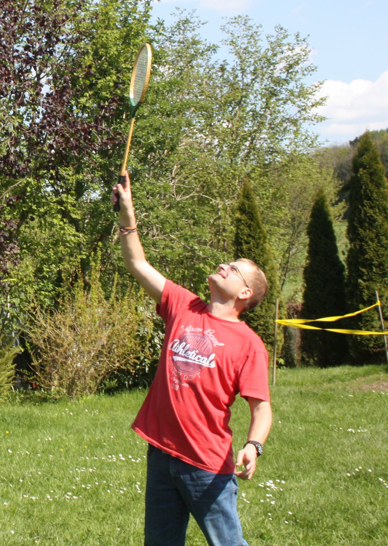 Ein Mann in einem roten T-Shirt hält einen Federballschläger in die Luft. Er wartet darauf, den Federball zu schlagen. Es ist sonnig und im Hintergrund sind Wiese und Bäume zu sehen.
